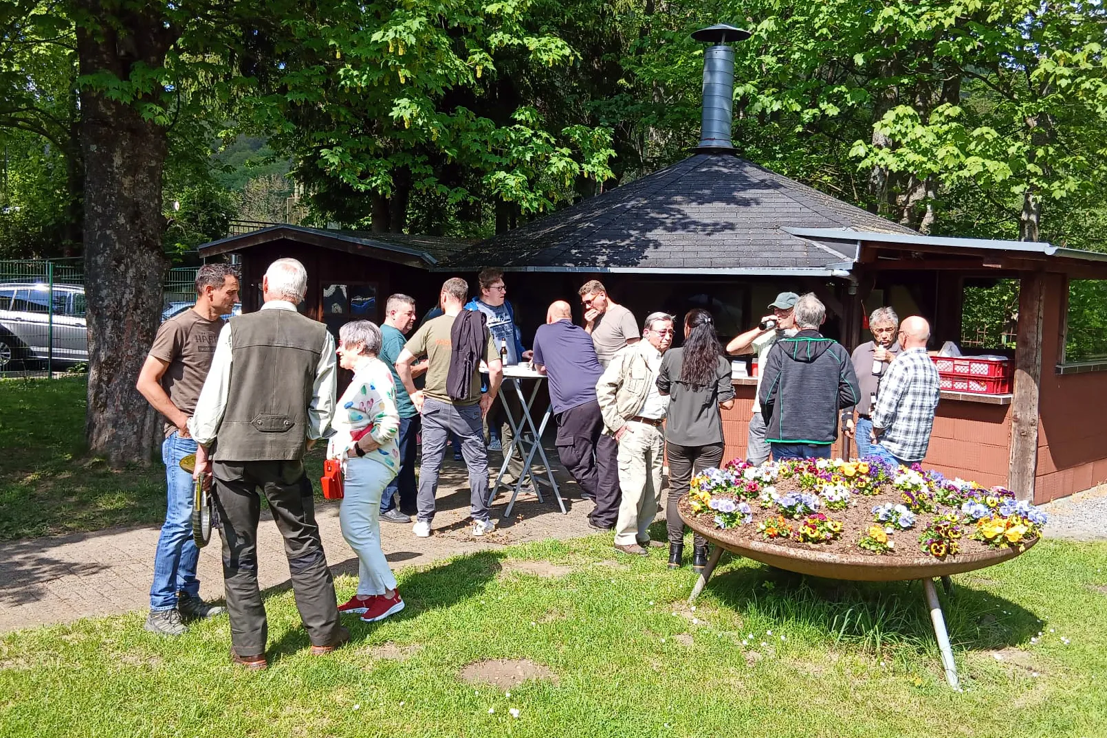 Menschen stehen vor einem Grillstand im Freien, einige unterhalten sich, andere warten auf Essen. Im Hintergrund Bäume und ein Auto.