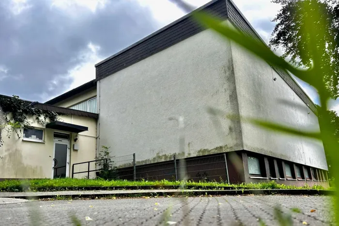 Ein großes, rechteckiges Gebäude mit einem dunklen Satteldach und einer hellen Fassade. Vor dem Gebäude verläuft ein Bürgersteig, dahinter ein kleiner Garten und ein Zaun.