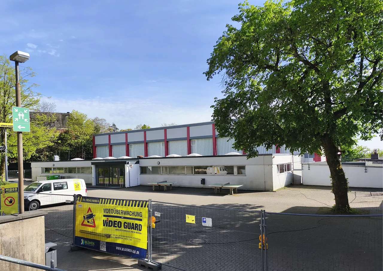 Ein helles, längliches Gebäude mit Flachdach und einer Reihe von Fenstern, davor ein Parkplatz mit einem weißen Lieferwagen und eine Absperrung mit Hinweisschildern zur Videoüberwachung.