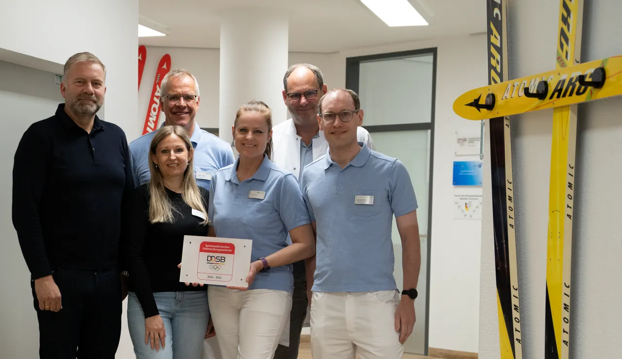 Fünf Personen stehen in einem hellen Raum vor einer Reihe von Skiern. Sie halten ein Schild mit dem Logo des Deutschen Olympischen Sportbundes (DOSB) und dem Gültigkeitszeitraum 2026–2034.