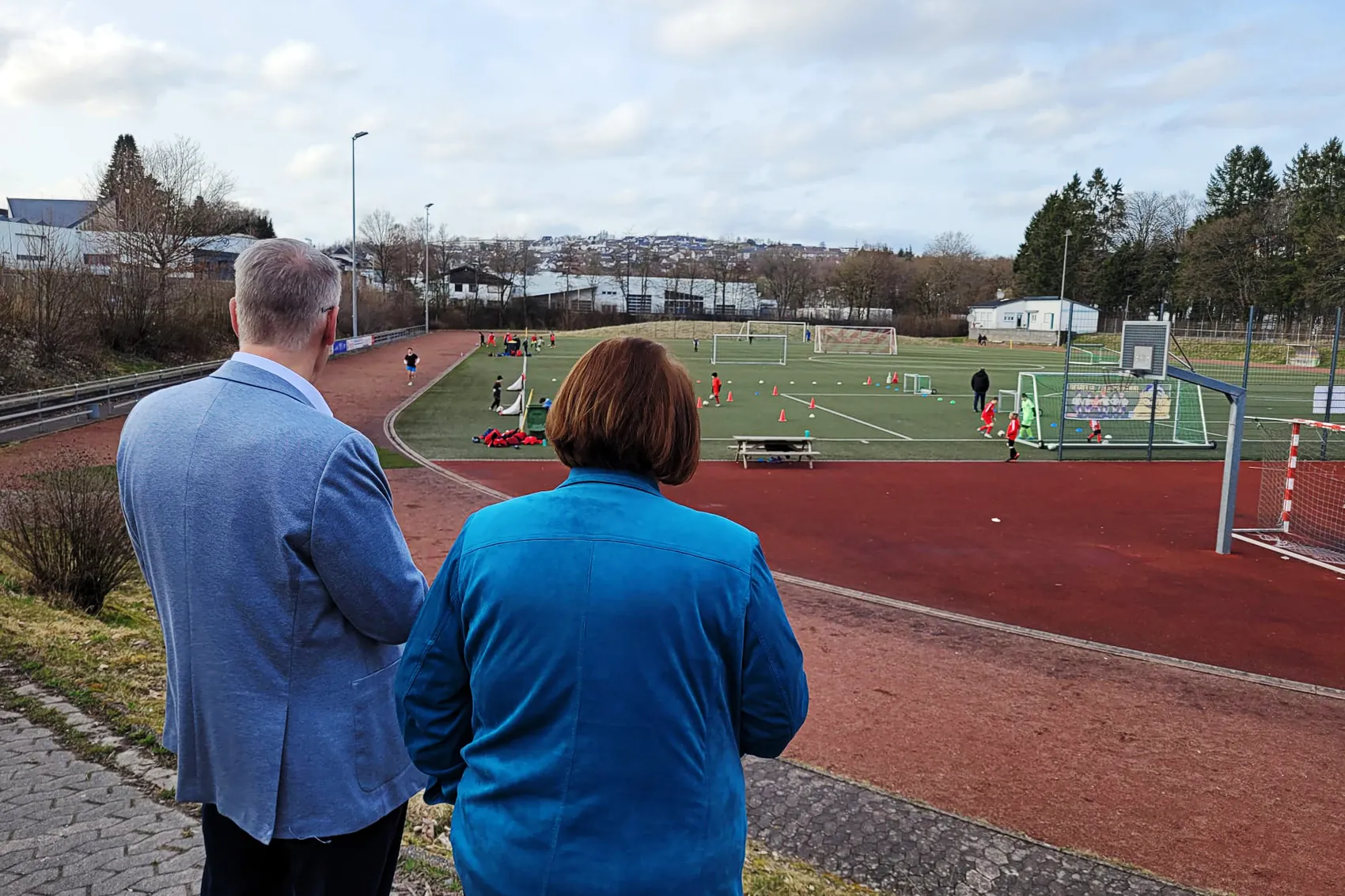 Zwei Personen mit dem Rücken zum Betrachter blicken auf einen Fußballplatz mit trainierenden Kindern. Im Hintergrund eine Stadtlandschaft und Bäume.