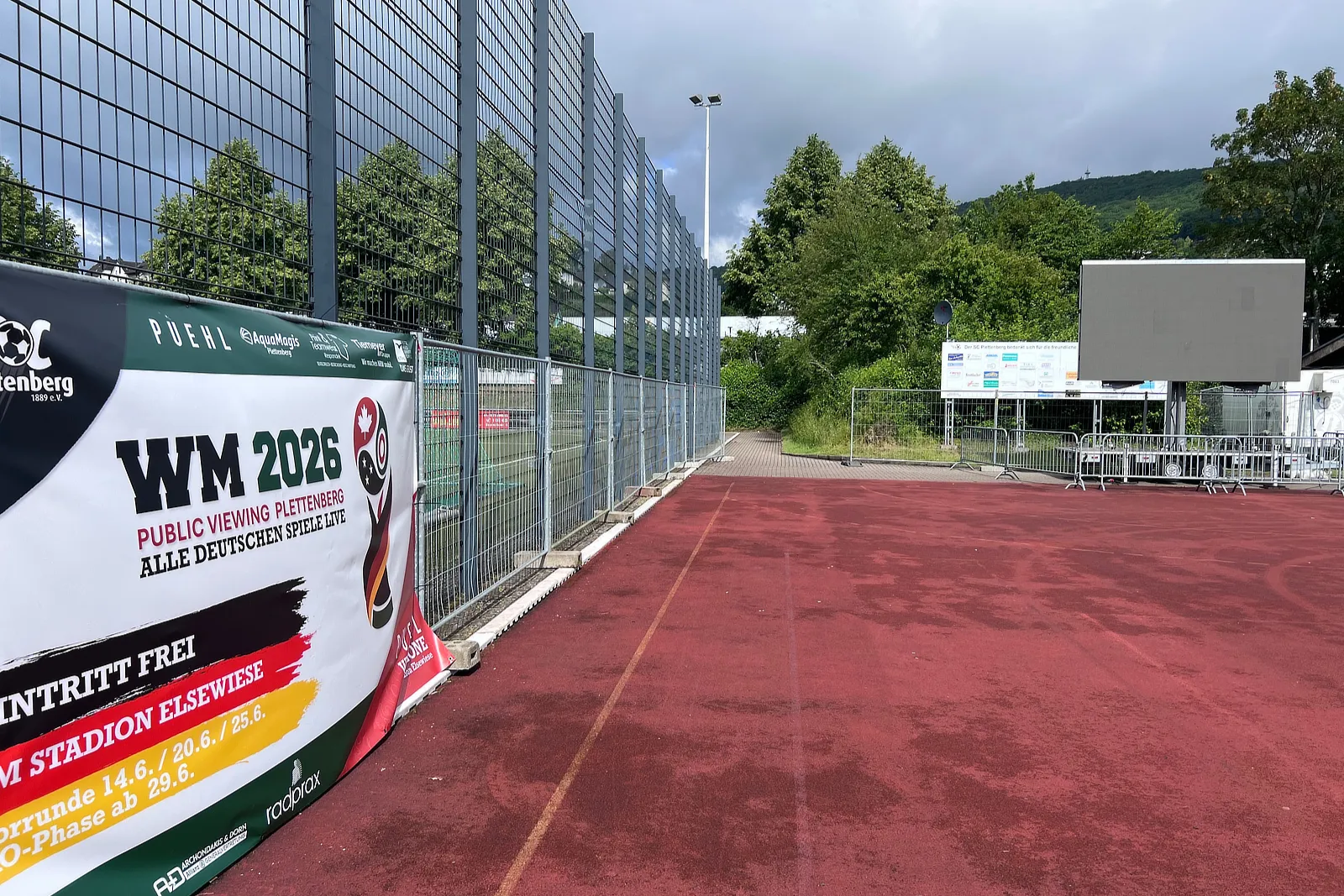 Ein Banner kündigt das Public Viewing der WM 2026 im Stadion Elsewiese in Plettenberg an. Im Vordergrund ist ein roter Sportplatz zu sehen, der durch eine blaue Bande begrenzt wird.