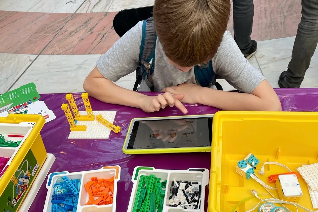 Ein Kind tippt auf ein Tablet, das eine Bauanleitung für ein Lego-Modell zeigt. Vor ihm liegen verschiedene Lego-Teile in farbigen Sortierboxen.