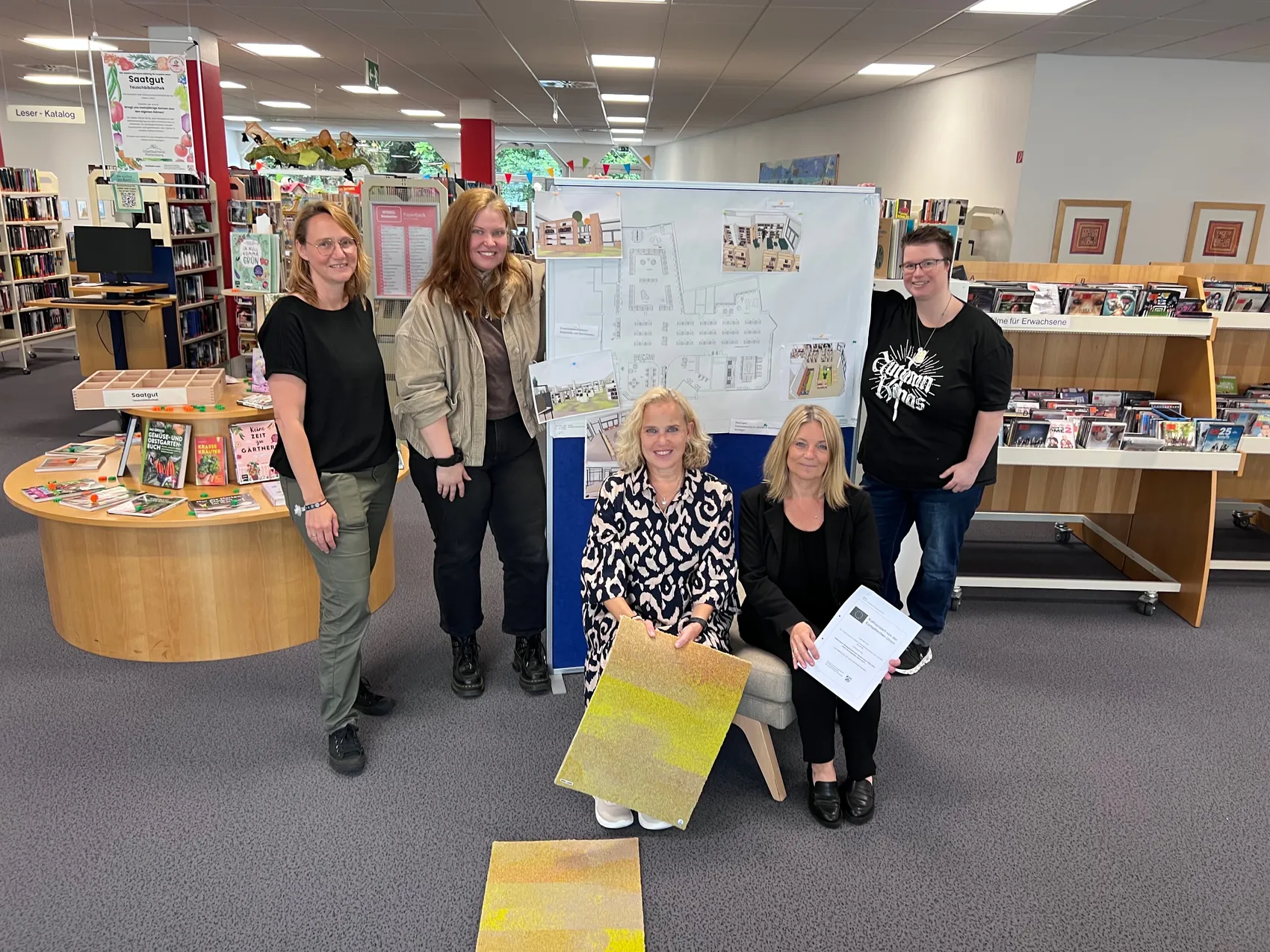 Fünf Frauen stehen in einer Bibliothek vor einer Skizze des neuen Raumlayouts. Sie halten Dokumente und lächeln in die Kamera.