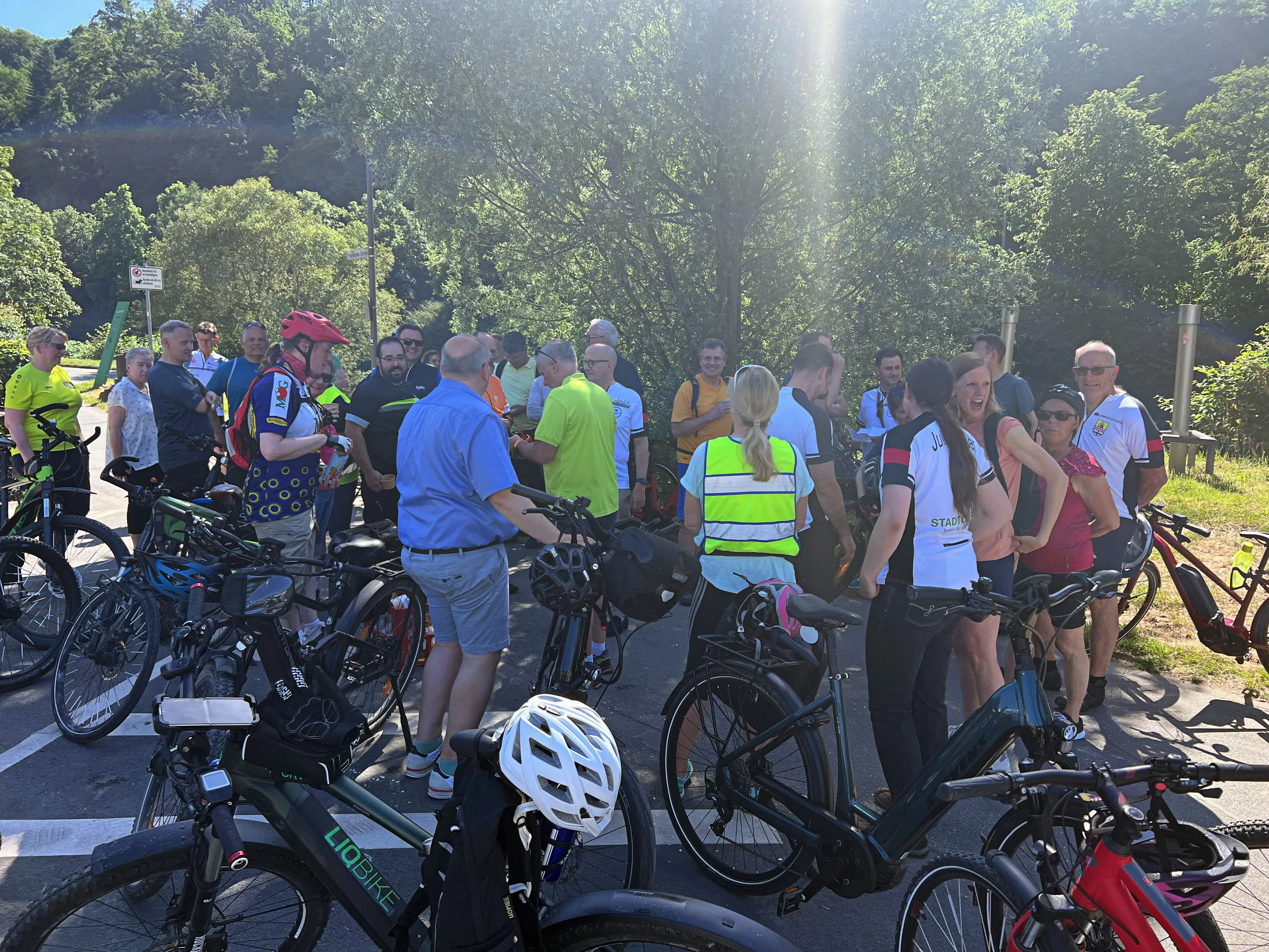 Eine Gruppe Radfahrer steht und unterhält sich auf einem Parkplatz, umgeben von Fahrrädern. Im Hintergrund sind Bäume und ein Verkehrsschild zu sehen.