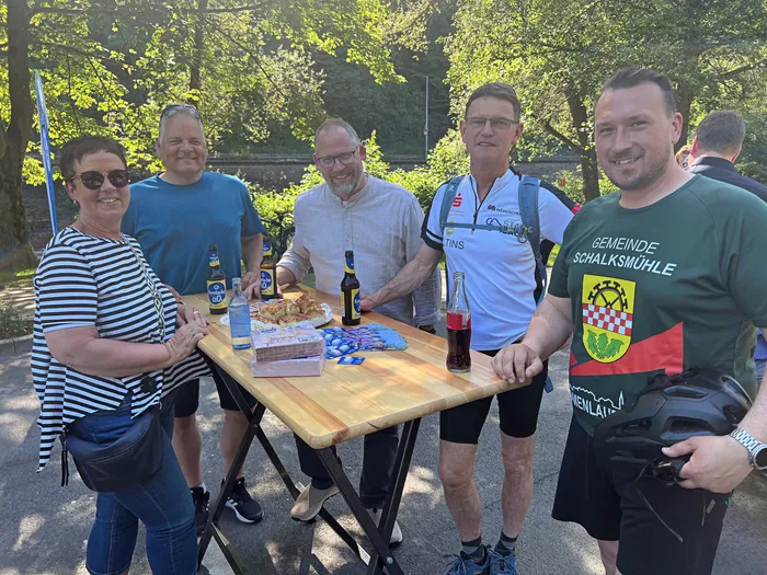 Eine Gruppe von sechs Männern und einer Frau steht lächelnd um einen Tisch mit Getränken und Snacks. Ein Mann trägt ein T-Shirt mit dem Wappen der Gemeinde Schalksmühle.