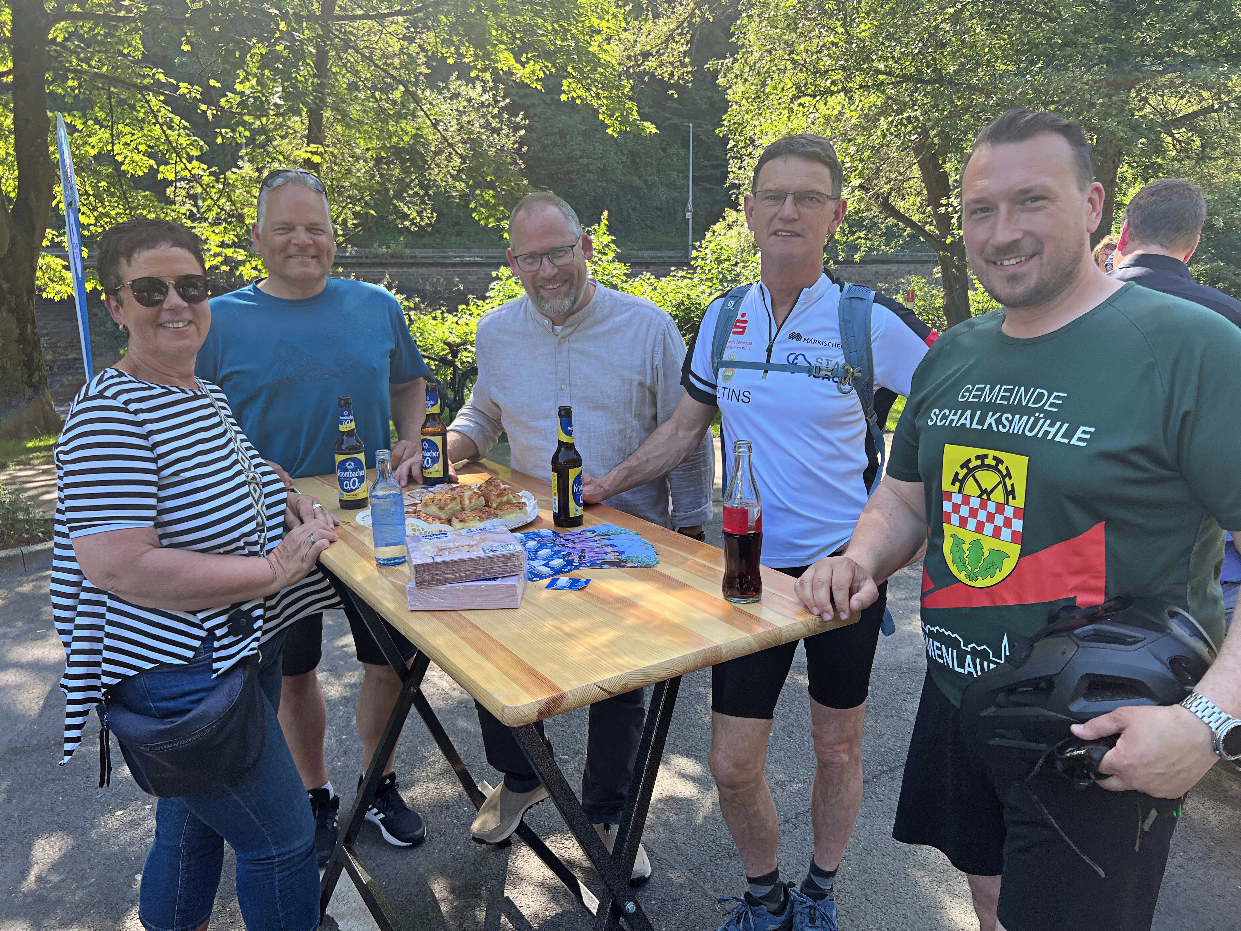Eine Gruppe von sechs Männern und einer Frau steht lächelnd um einen Tisch mit Getränken und Snacks. Ein Mann trägt ein T-Shirt mit dem Wappen der Gemeinde Schalksmühle.