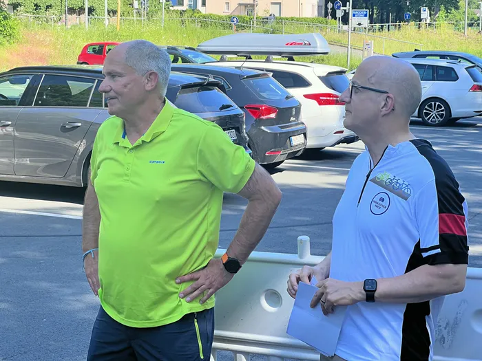 Zwei Männer stehen auf einem Parkplatz vor geparkten Autos. Der Mann links trägt ein hellgrünes Poloshirt, der Mann rechts ein weißes T-Shirt mit einem Fahrrad-Logo.