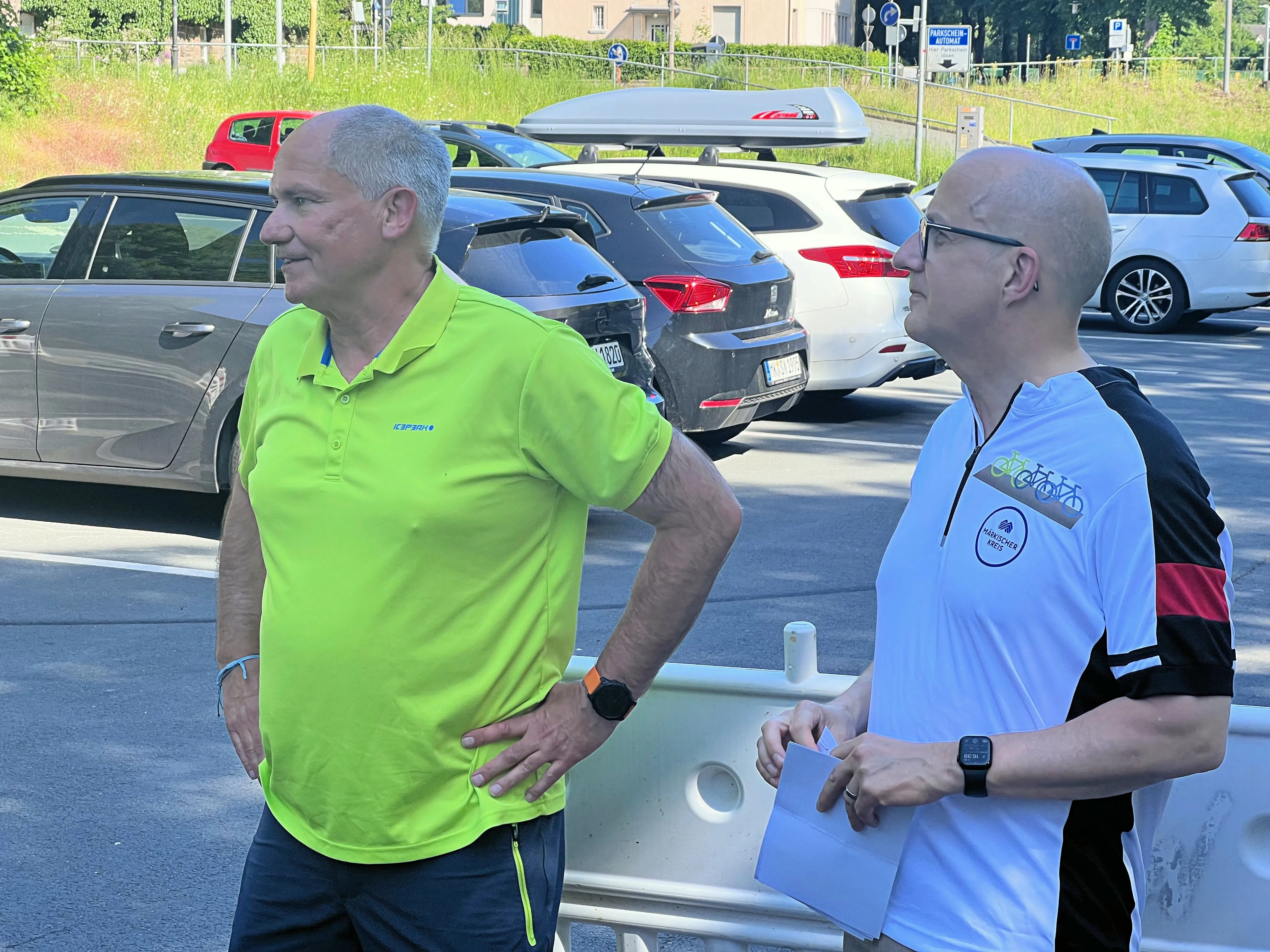 Zwei Männer stehen auf einem Parkplatz vor geparkten Autos. Der Mann links trägt ein hellgrünes Poloshirt, der Mann rechts ein weißes T-Shirt mit einem Fahrrad-Logo.