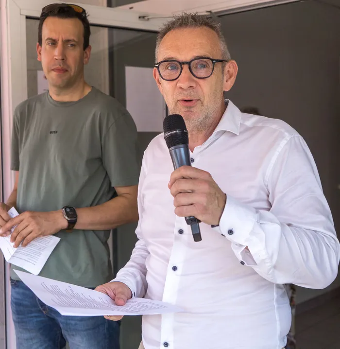 Ein Mann mit Brille spricht in ein Handmikrofon, während er Notizen hält. Im Hintergrund steht ein weiterer Mann.