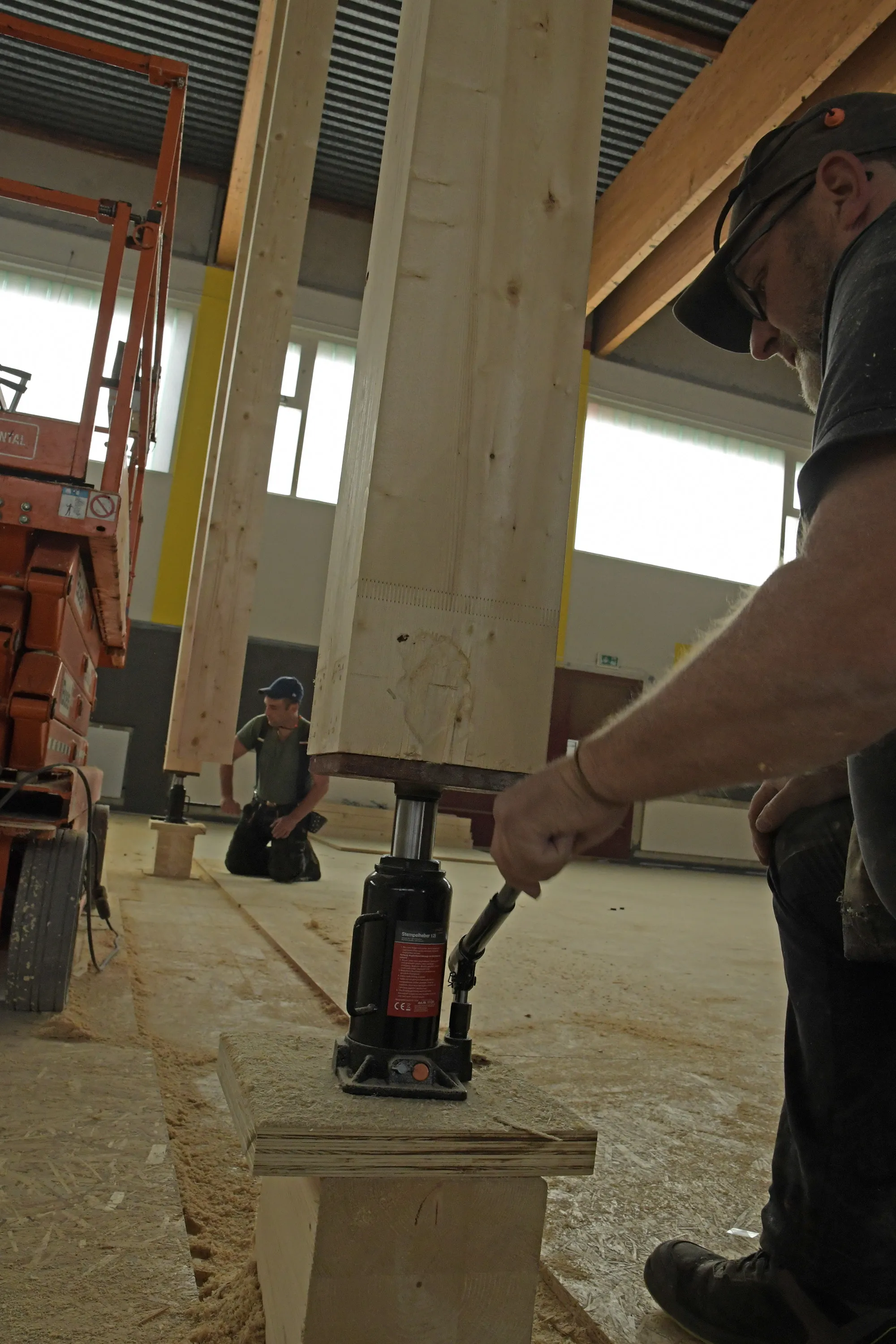 Ein Bauarbeiter bedient einen Stempelheber, um einen großen Holzbinder anzuheben. Im Hintergrund ist ein weiterer Arbeiter zu sehen und ein Gabelstapler.