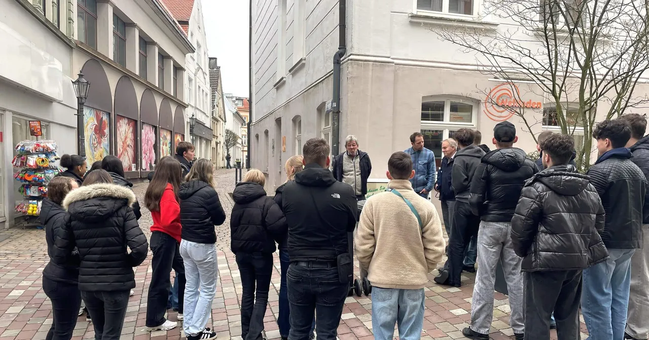 Eine Gruppe Jugendlicher steht auf einem Kopfsteinpflasterweg in einer Stadt und lauscht einer Frau, die ihnen etwas erklärt. Im Hintergrund sind historische Gebäude und ein Geschäft zu sehen.
