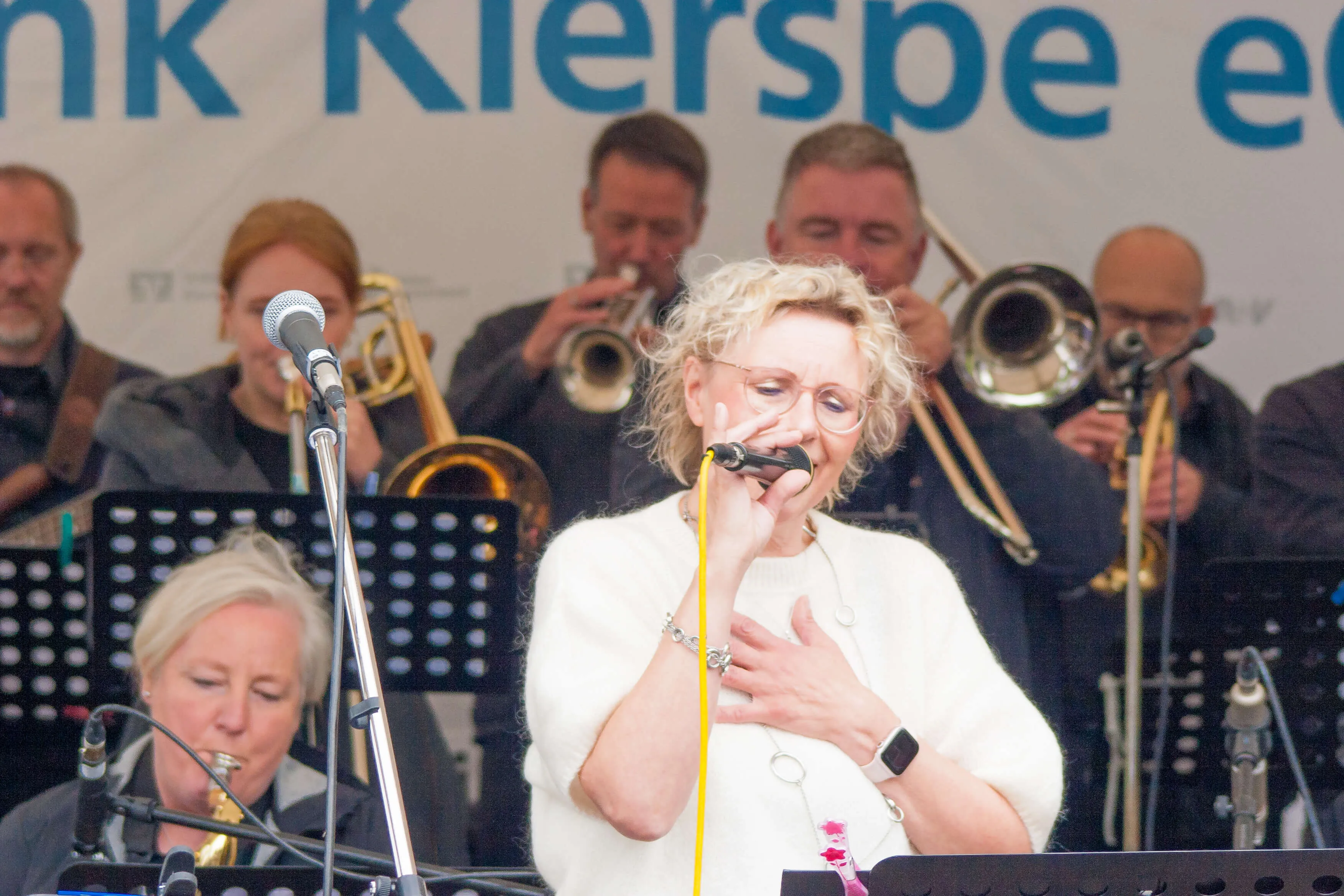 Sängerin mit gelocktem Haar und Brille singt in ein Mikrofon, umgeben von Musikern einer Bigband mit Blechblasinstrumenten.