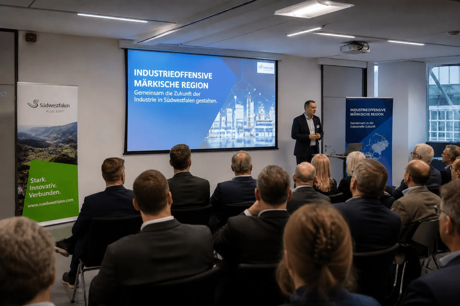 Ein Mann hält vor einem Publikum in einem Konferenzraum einen Vortrag, während im Hintergrund eine Präsentation mit dem Titel „Industrieoffensive Märkische Region“ zu sehen ist.