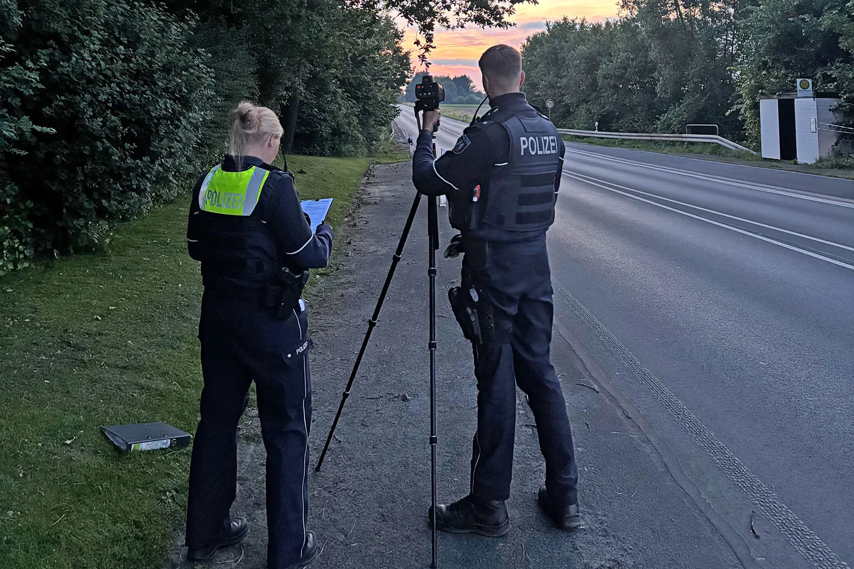 Zwei Polizisten sichern eine Geschwindigkeitsmessung mit einem Lasergerät an einer Landstraße. Einer notiert Daten, während der andere das Gerät bedient.
