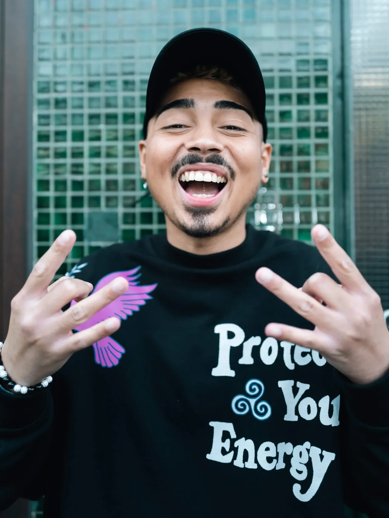 Max Kopp lacht mit offenem Mund und zeigt mit beiden Händen das Rocker-Handzeichen. Er trägt ein schwarzes Sweatshirt mit der Aufschrift „Protect Your Energy“.