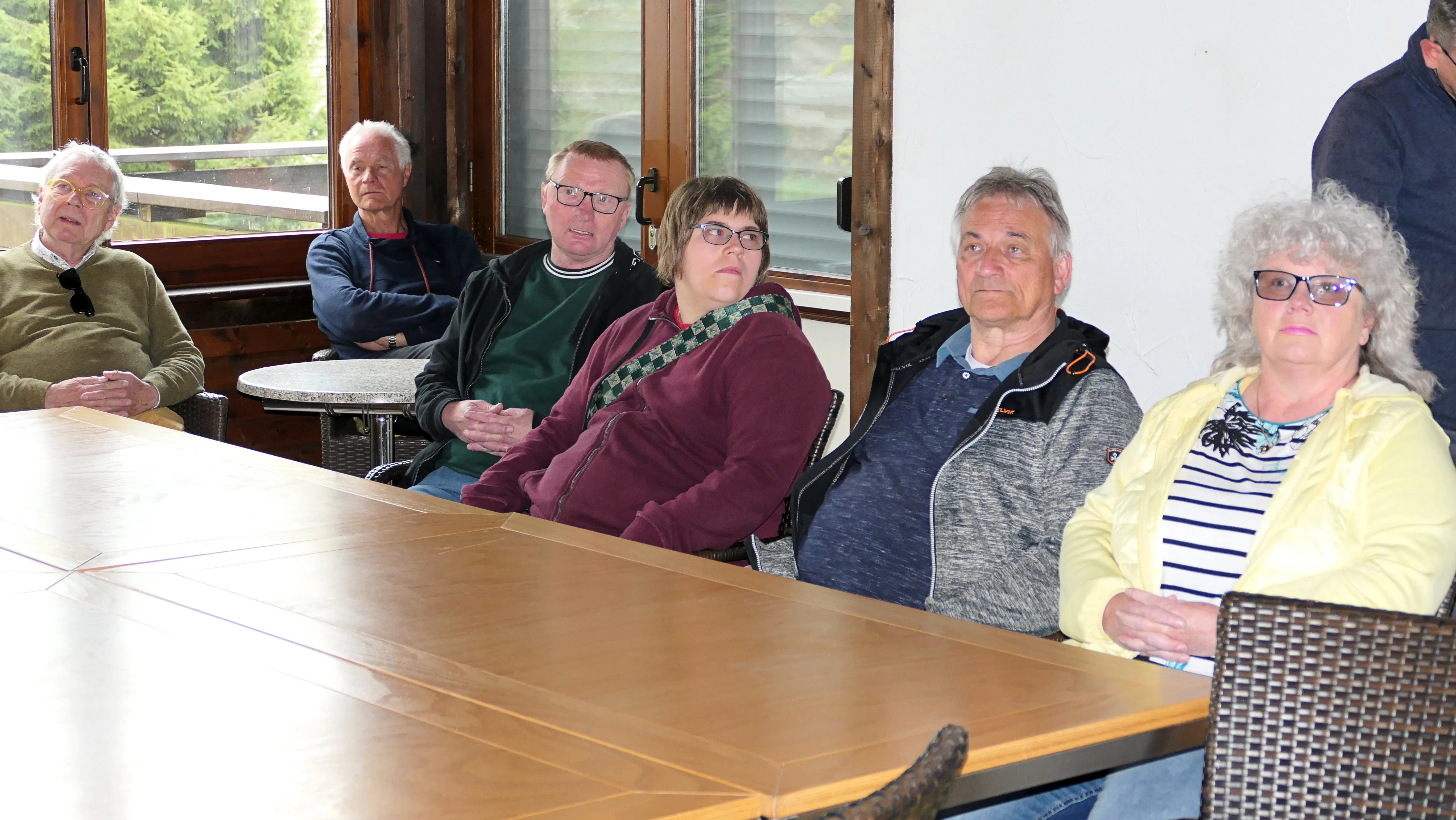 Sechs ältere Menschen sitzen an einem großen Tisch in einem hellen Raum und schauen nach vorn.