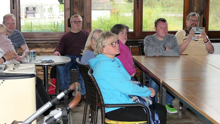 Eine Gruppe von sechs Personen sitzt an einem langen Tisch in einem hellen Raum mit Fenstern im Hintergrund und verfolgt aufmerksam eine Aktion, möglicherweise ein Pickleball-Spiel.