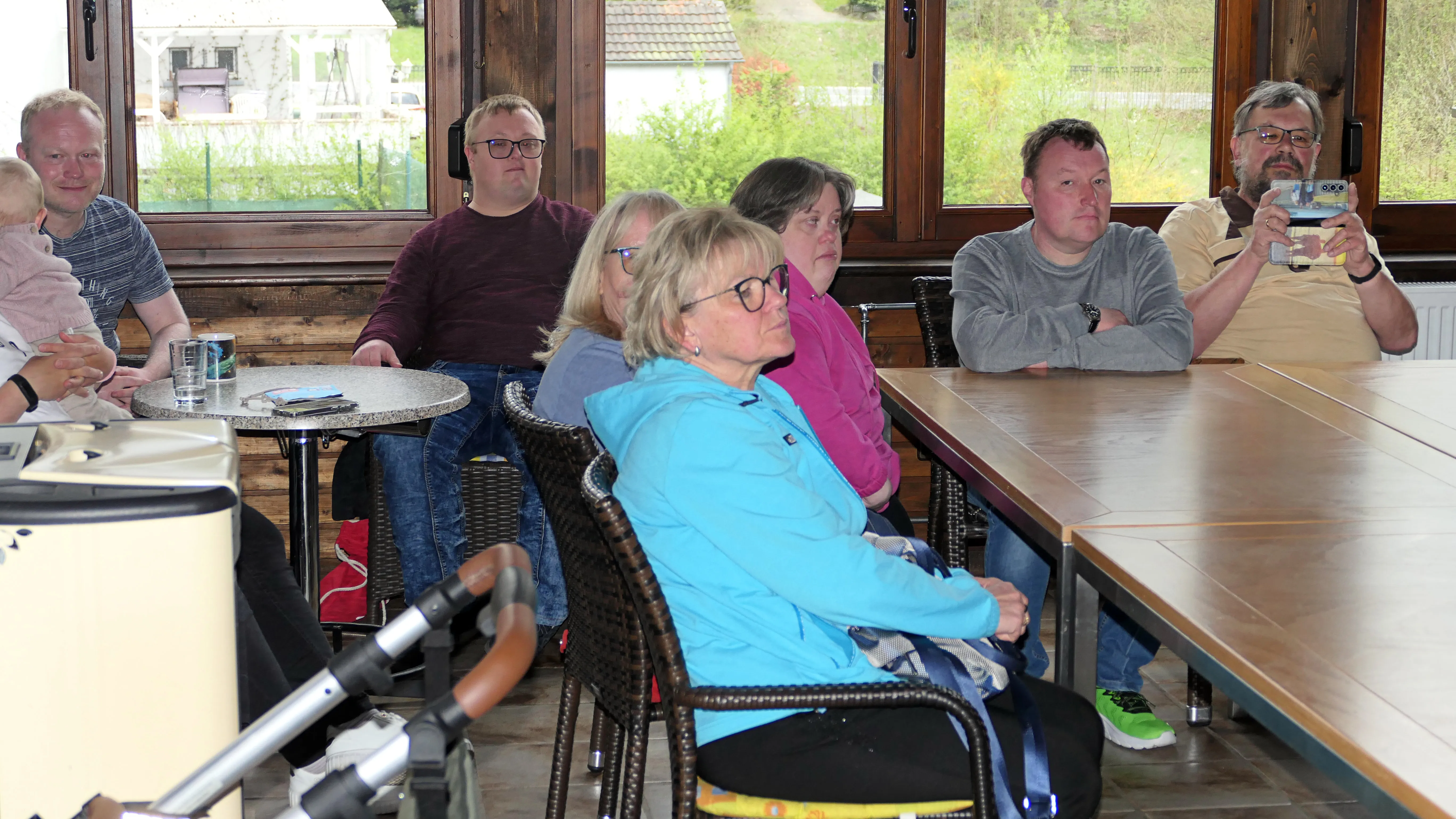 Eine Gruppe von sechs Personen sitzt an einem langen Tisch in einem hellen Raum mit Fenstern im Hintergrund und verfolgt aufmerksam eine Aktion, möglicherweise ein Pickleball-Spiel.