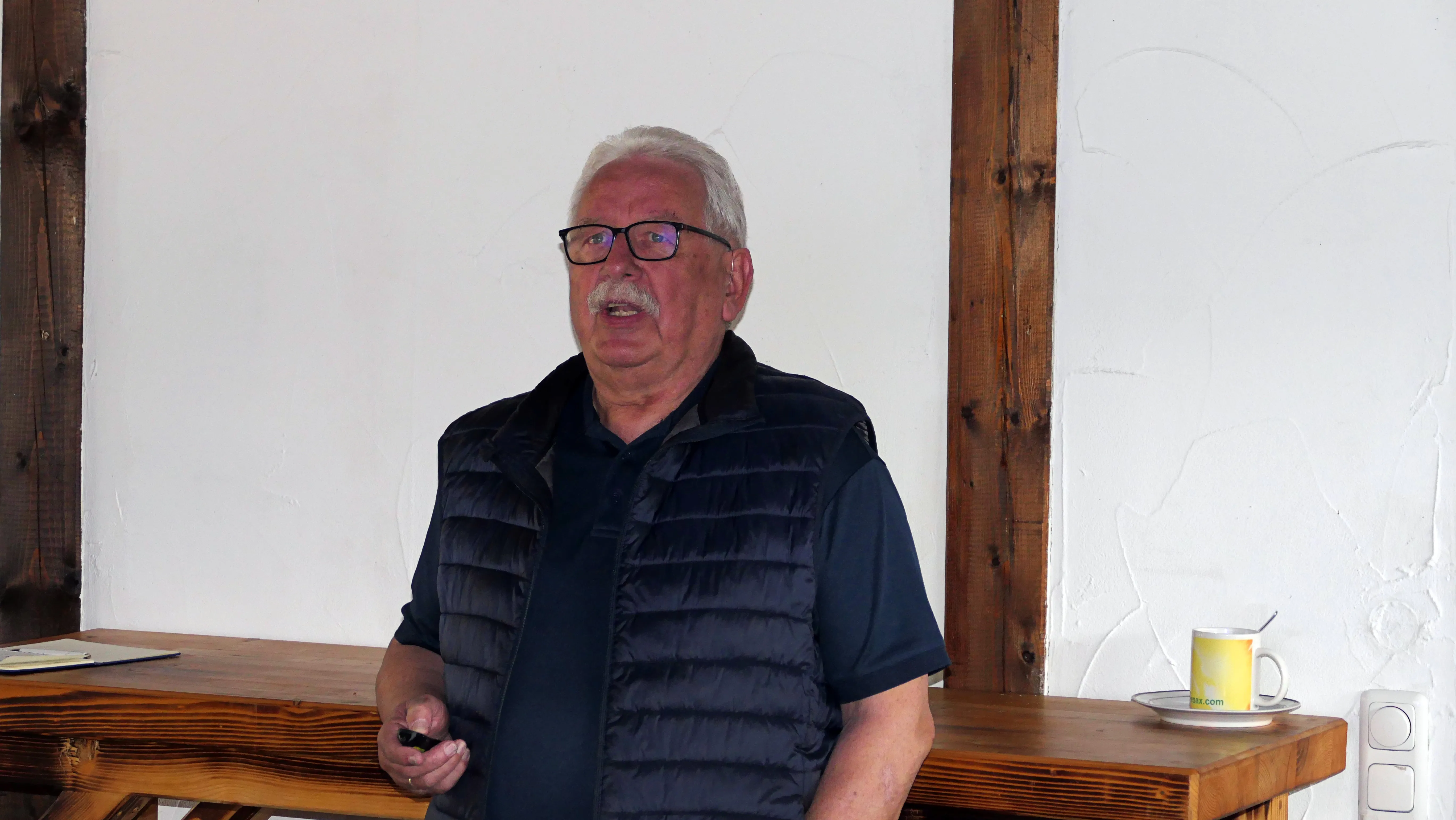 Ein älterer Mann mit Brille und grauem Haar spricht in die Kamera, während er an einem Tisch sitzt. Neben ihm steht eine gelbe Tasse mit Aufschrift.