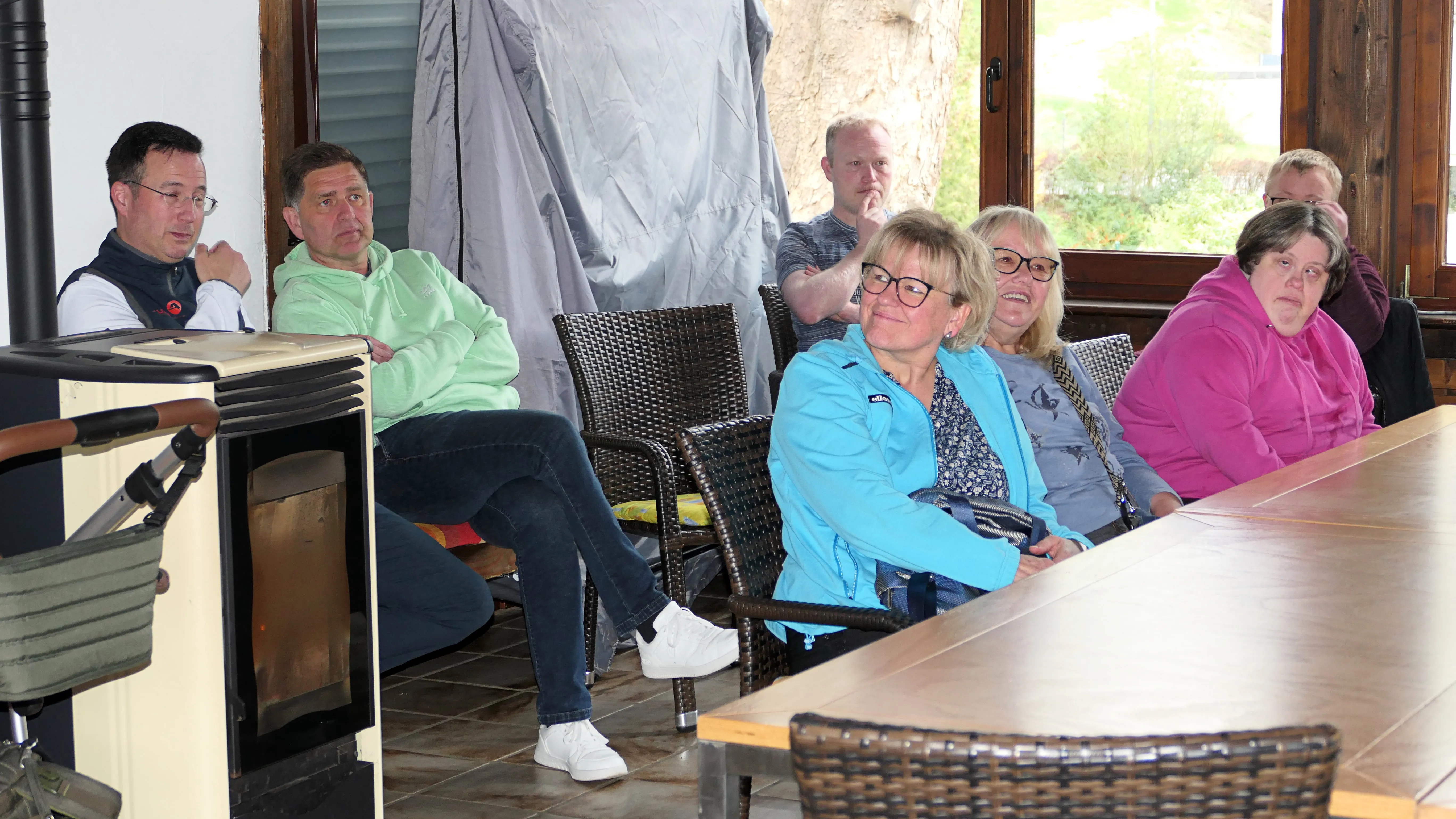Eine Gruppe von sieben Personen sitzt an einem großen Holztisch und schaut in eine Richtung, möglicherweise auf ein Pickleball-Feld. Im Vordergrund eine Frau mit Brille in hellblauer Jacke und weiße Sneaker.