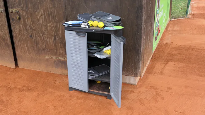 Grauer Schrank mit offen stehender Tür, darin befinden sich Pickleball-Schläger, Bälle und Taschen auf einer roten Oberfläche. Im Hintergrund ist ein roter Platz zu sehen.