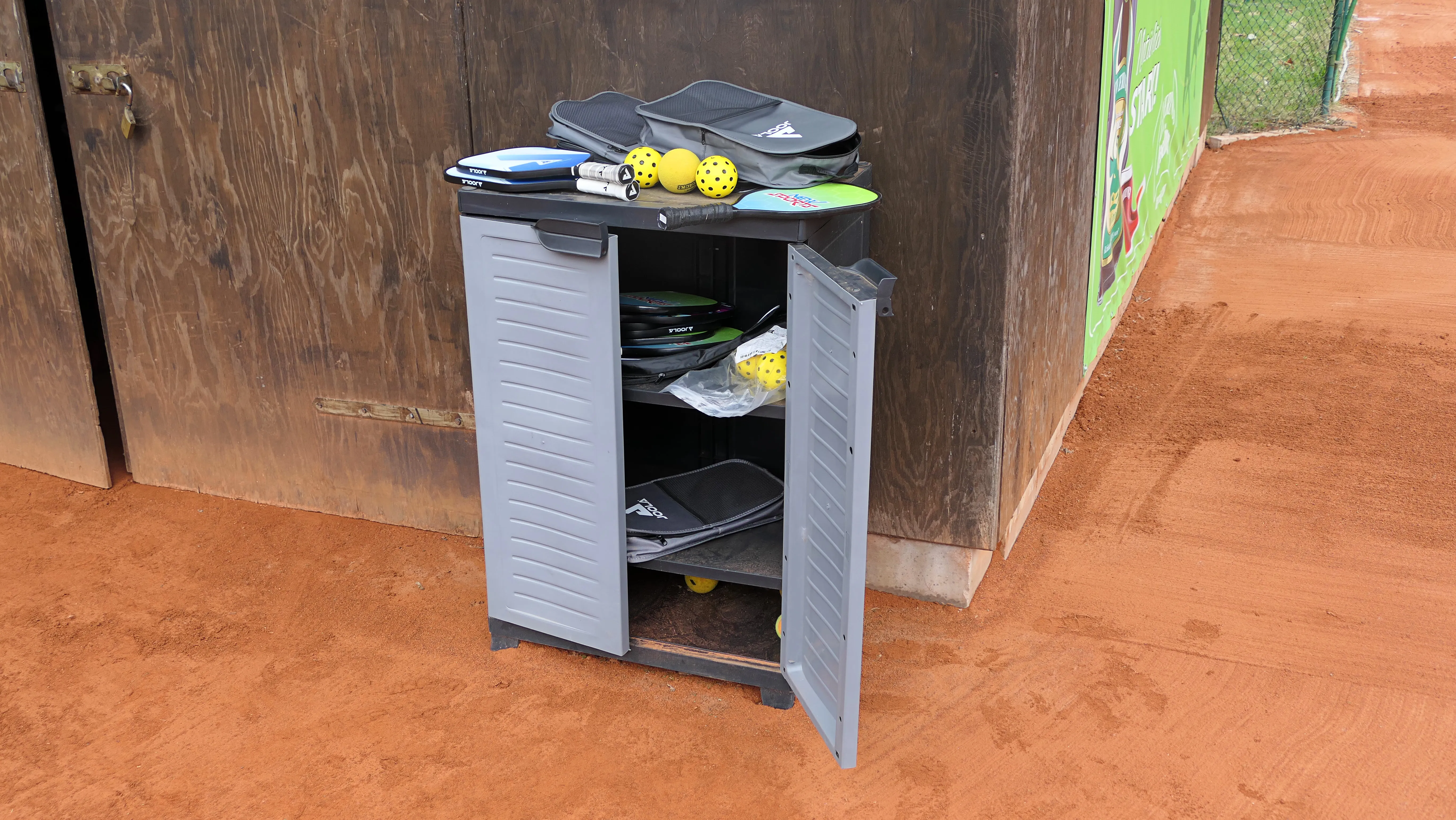 Grauer Schrank mit offen stehender Tür, darin befinden sich Pickleball-Schläger, Bälle und Taschen auf einer roten Oberfläche. Im Hintergrund ist ein roter Platz zu sehen.