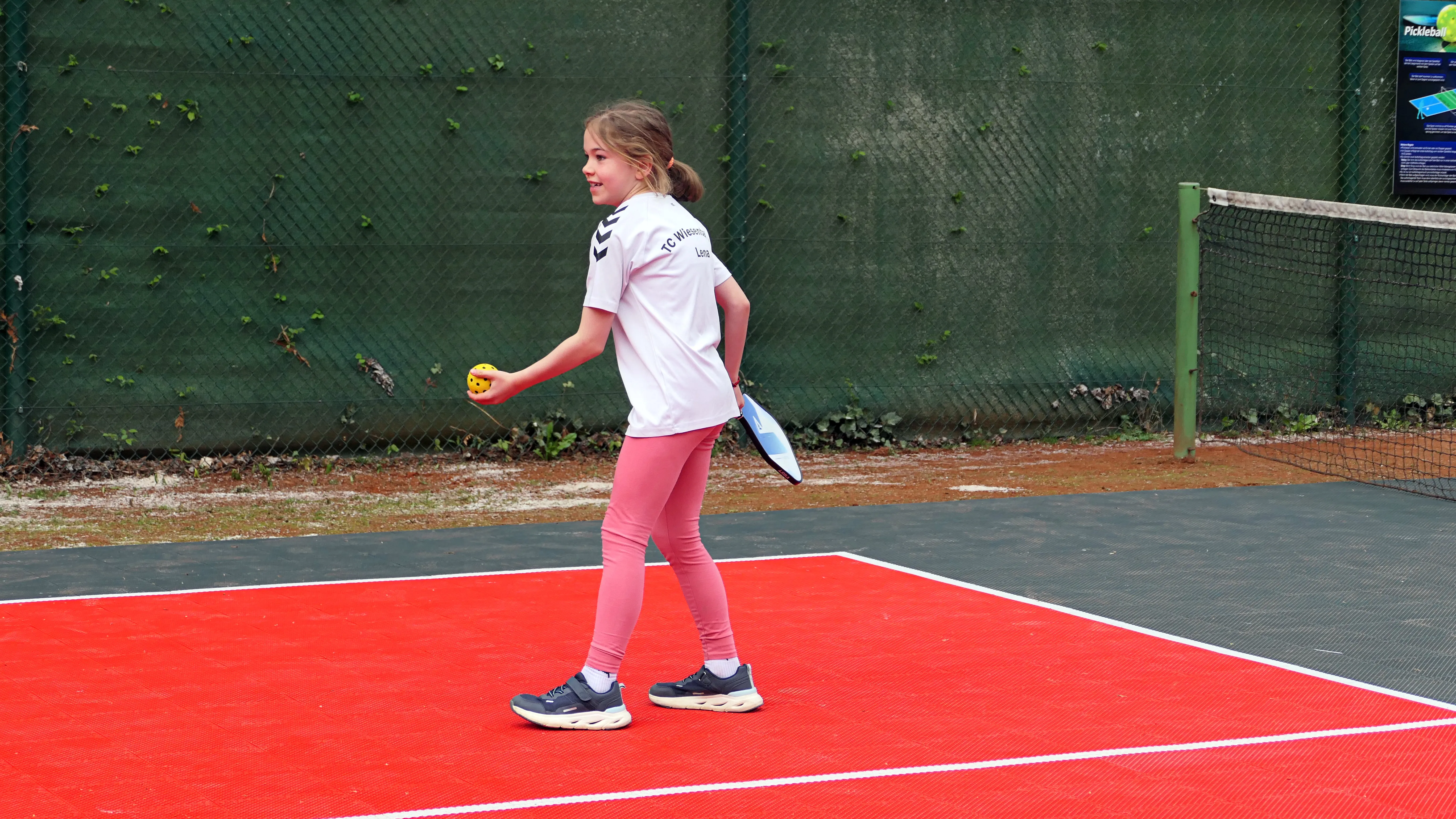 Ein junges Mädchen hält einen gelben Pickleball und einen Schläger bereit, um auf einem roten Pickleballfeld zu spielen. Im Hintergrund ein grüner Zaun und ein Netz.