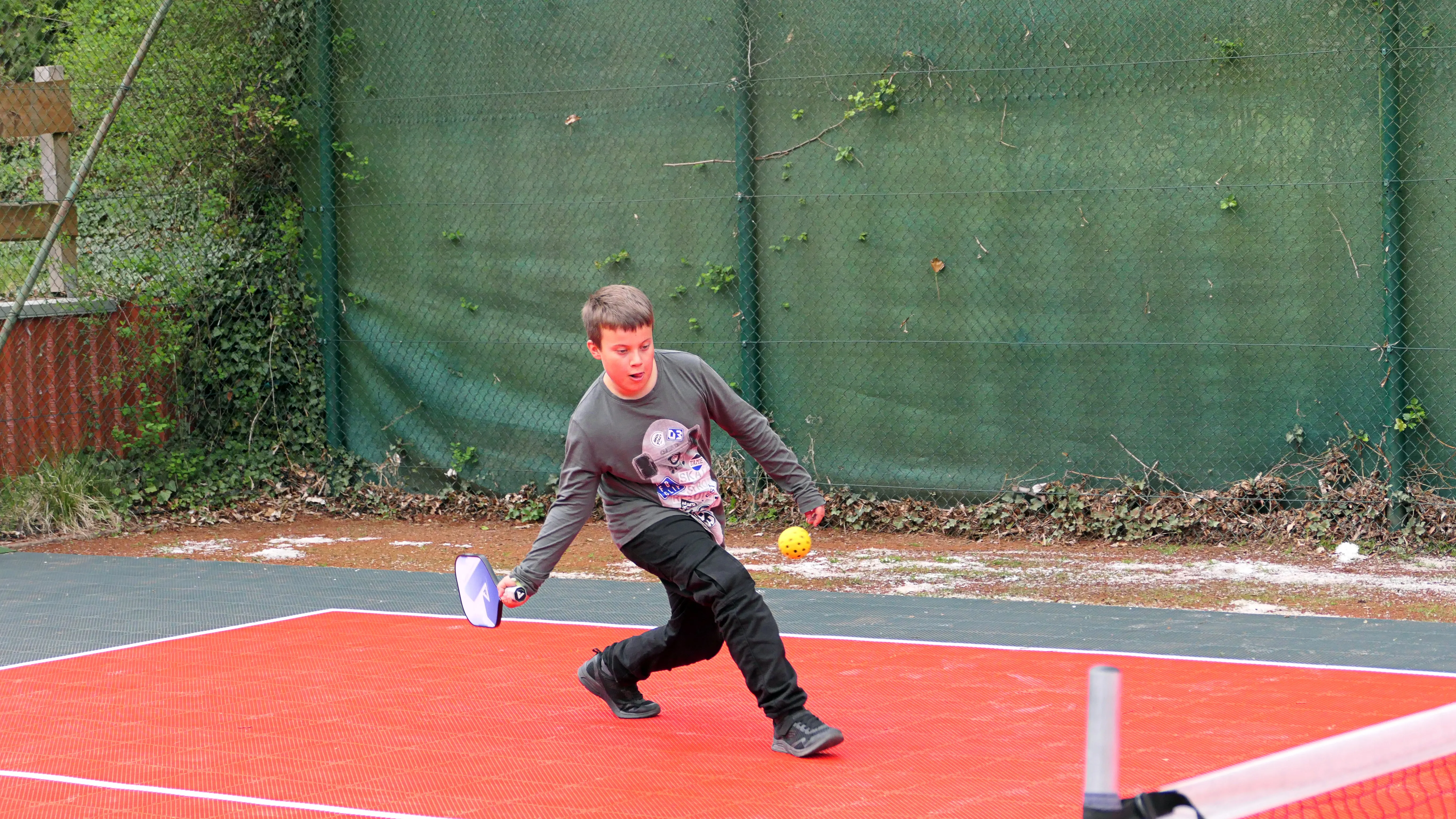 Junge schwingt Schläger beim Pickleball auf rotem Spielfeld, gelber Ball in der Luft.