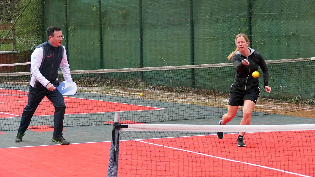 Zwei Personen spielen Pickleball auf einem roten Platz mit schwarzem Netz. Die Frau schlägt den Ball, der Mann bereitet sich auf die Rückgabe vor.