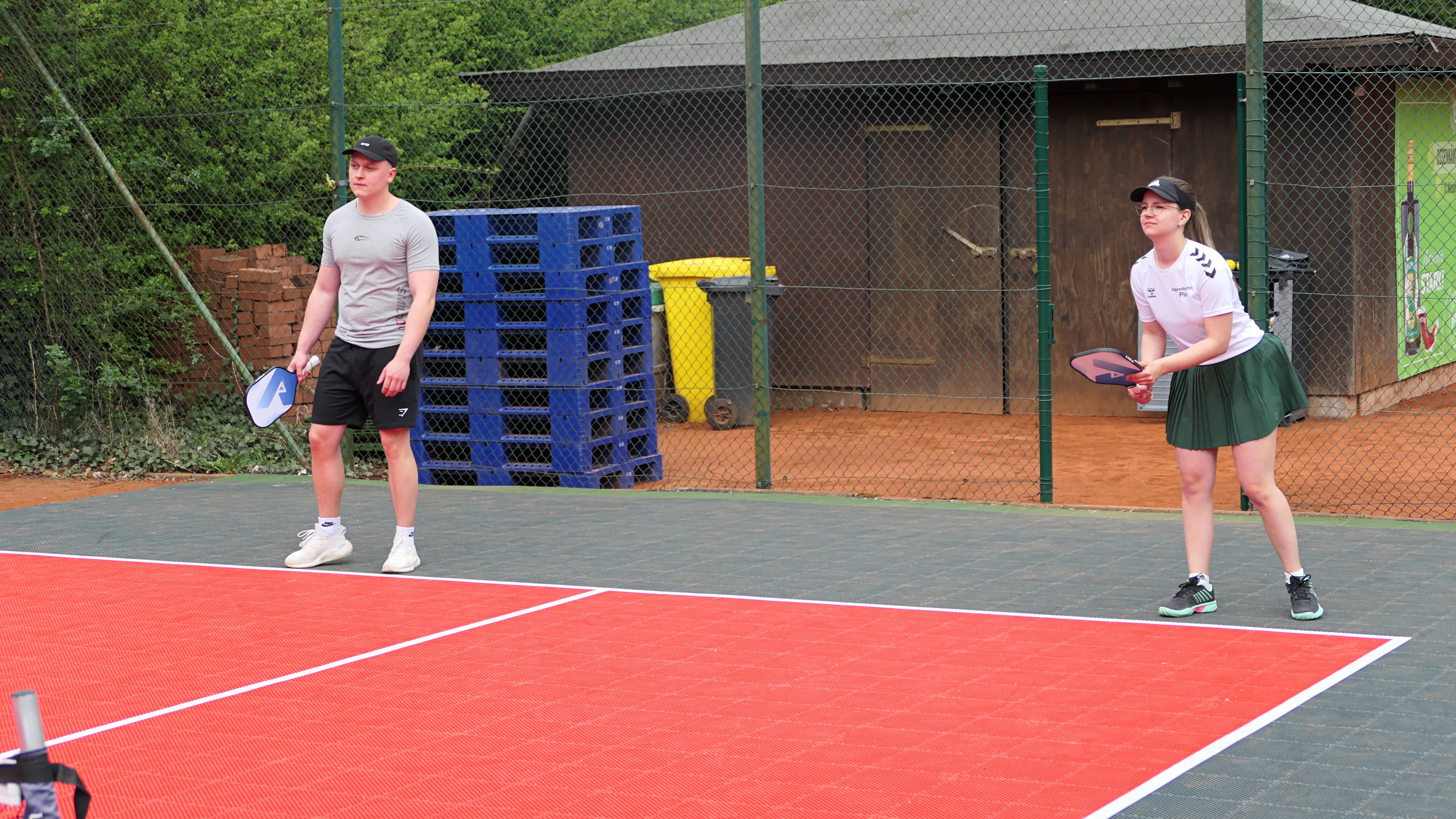 Zwei junge Erwachsene stehen auf einem roten Pickleball-Platz, bereit zum Spiel mit Schlägern und Ball.