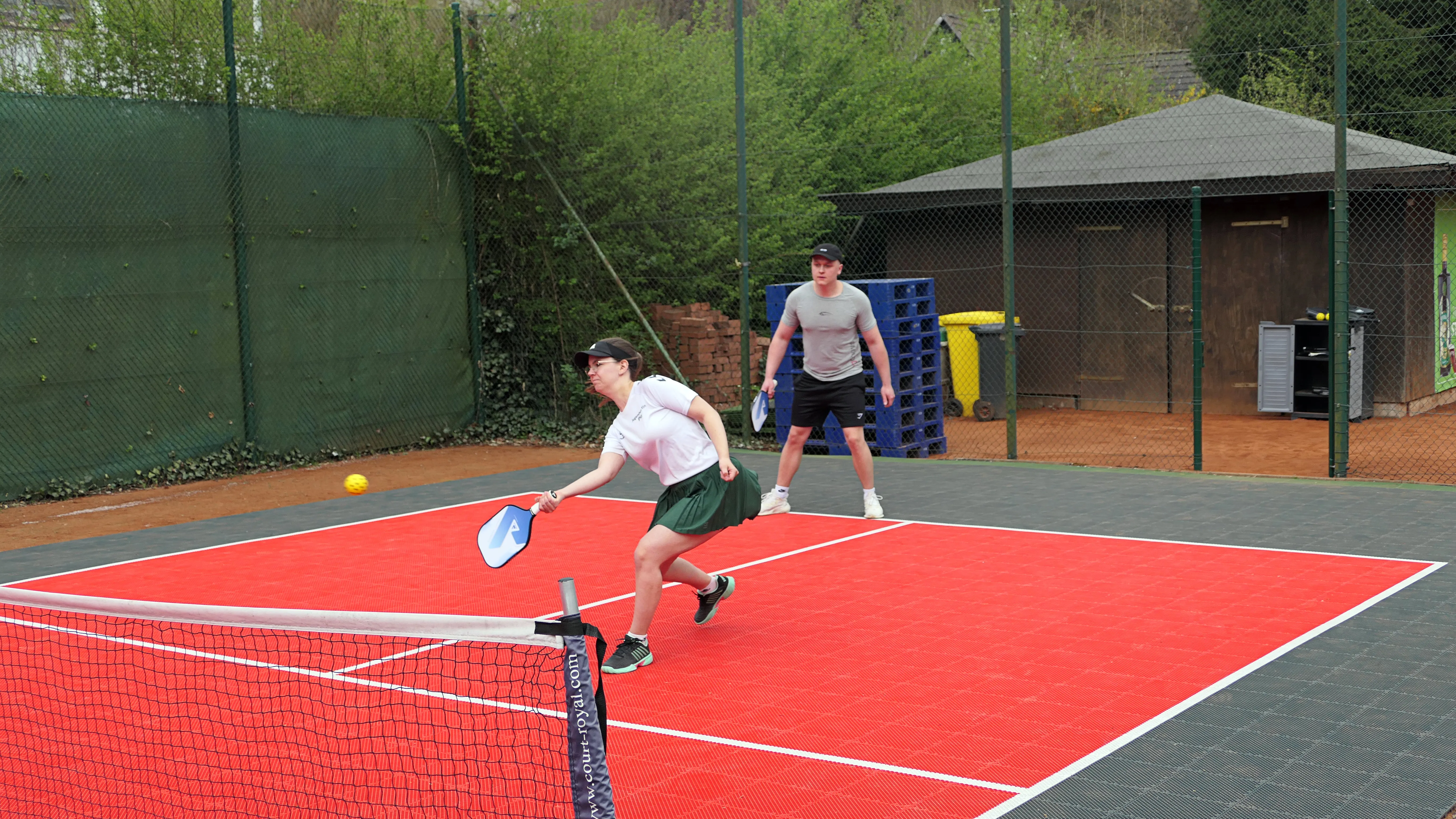 Zwei junge Erwachsene spielen Pickleball auf einem roten Platz mit schwarzem Netz, im Hintergrund ein grüner Garten und ein Unterstand.