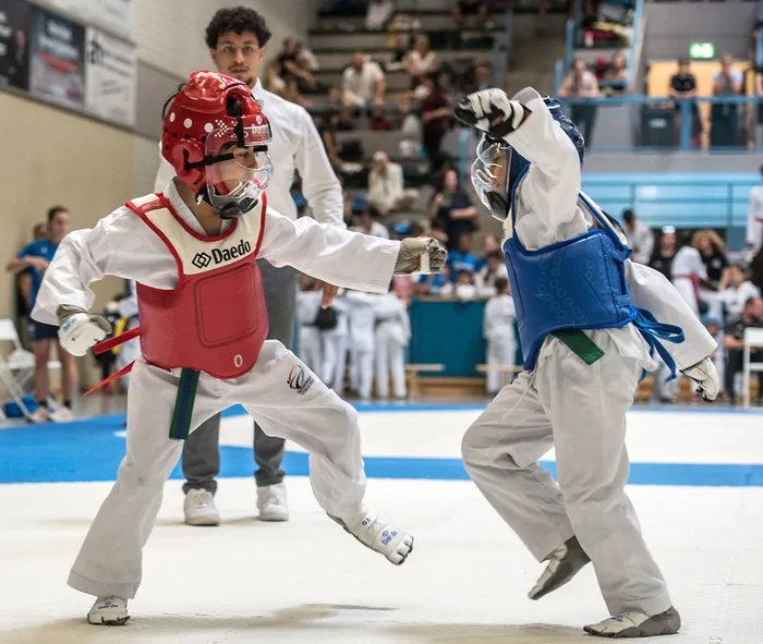 Zwei Taekwondo-Kämpfer in voller Montur stehen sich auf einer blauen Matte gegenüber, der eine trägt Rot, der andere Blau. Im Hintergrund sind Zuschauer und ein Trainer zu sehen.