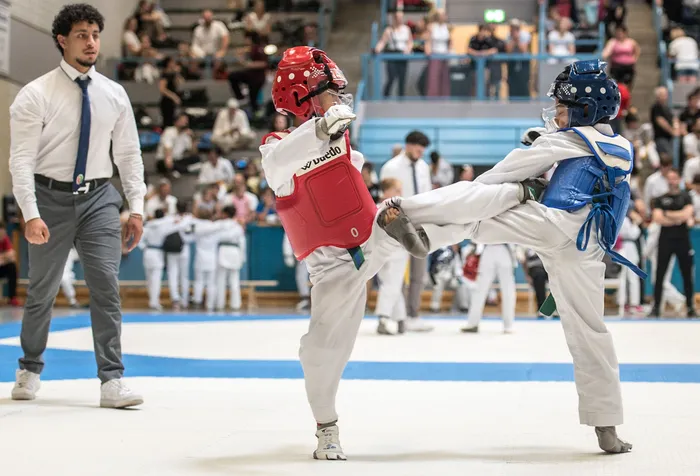 Zwei Taekwondo-Kämpfer in weißen Anzügen treten in einer Sporthalle gegeneinander an, während ein Trainer im Hintergrund zuschaut. Der Kämpfer links trägt rote Schutzausrüstung, der andere blaue.