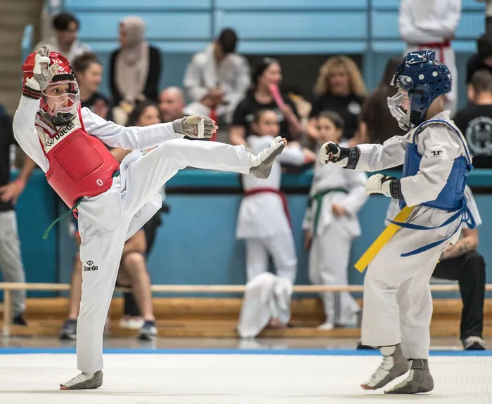 Zwei Taekwondo-Athleten kämpfen in einem Wettkampf. Der Athlet links trägt rote Schutzkleidung und führt einen Tritt aus, während der Athlet rechts in blauer Kleidung blockt. Im Hintergrund sind Zuschauer auf den Tribünen zu sehen.