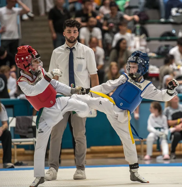 Zwei Taekwondo-Kämpfer in voller Montur treten gegeneinander an, während ein Schiedsrichter im Hintergrund aufmerksam beobachtet. Der Kämpfer links trägt rote Schutzkleidung, der rechts blaue.