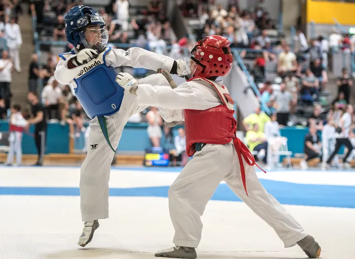 Zwei Taekwondo-Athleten in voller Montur kämpfen in einer Halle vor Zuschauern. Der Athlet links trägt einen blauen Brustpanzer, der Athlet rechts einen roten.