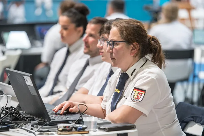 Eine Frau mit Brille und hochgestecktem Haar tippt auf einen Laptop. Im Hintergrund sind weitere Personen in weißer Kleidung zu sehen, vermutlich Teilnehmer oder Betreuer des Taekwondo-Turniers.