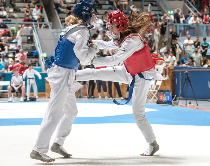 Zwei Taekwondo-Athleten in weißen Anzügen kämpfen in einem Wettkampf. Die Athletin in roter Ausrüstung führt einen Tritt aus, während ihre Gegnerin in blauem Schutz sich verteidigt. Im Hintergrund sind Zuschauer und die Sporthalle zu sehen.