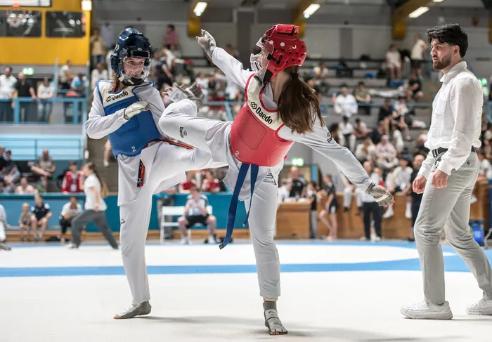 Zwei Taekwondo-Athletinnen in blauer und roter Schutzausrüstung kämpfen in einer Sporthalle vor Zuschauern. Ein Kampfrichter steht rechts im Bild.