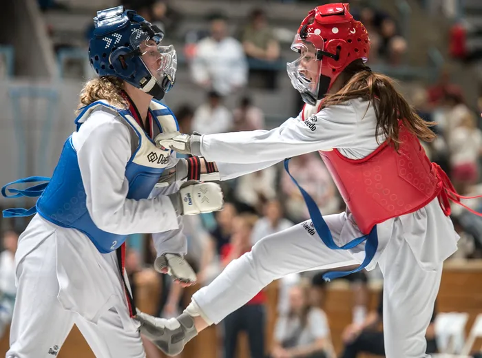 Zwei Taekwondo-Athletinnen in voller Montur kämpfen in einem Wettkampf. Die Sportlerin links trägt Blau, die rechts Rot.