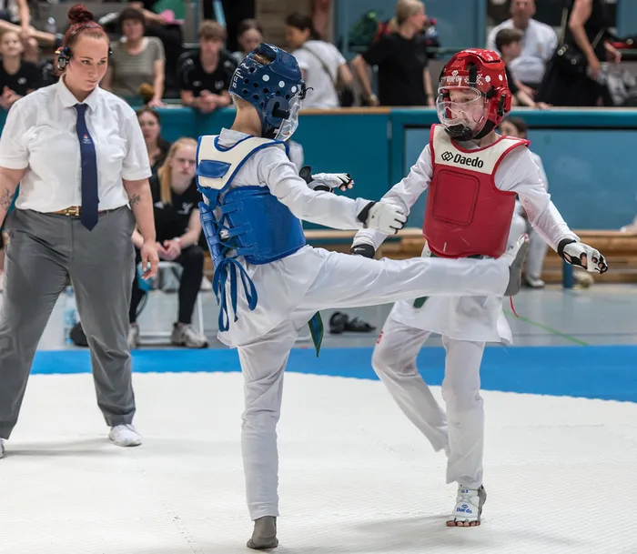 Zwei Taekwondo-Kämpfer in voller Schutzausrüstung treten in einem Wettkampf gegeneinander an, während eine Schiedsrichterin im Hintergrund aufmerksam beobachtet.