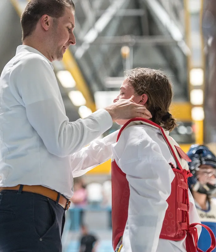 Ein Mann mit Hemd und Jeans justiert einem jungen Sportler mit roter Schutzausrüstung den Kopfschutz. Im Hintergrund sind weitere Sportler und eine große Halle zu sehen.