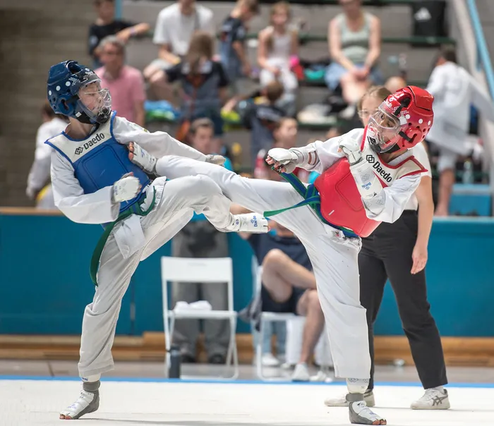 Zwei Taekwondo-Athleten in blauen und roten Schutzwesten kämpfen in einer Halle. Der Athlet in Blau führt einen Tritt aus, während der Athlet in Rot blockt. Im Hintergrund sind Zuschauer auf den Tribünen zu sehen.