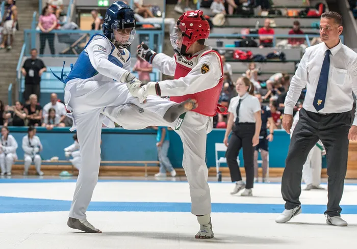 Zwei Taekwondo-Athleten kämpfen in einem Wettkampf, einer tritt, der andere blockt. Im Hintergrund stehen Zuschauer und Kampfrichter.