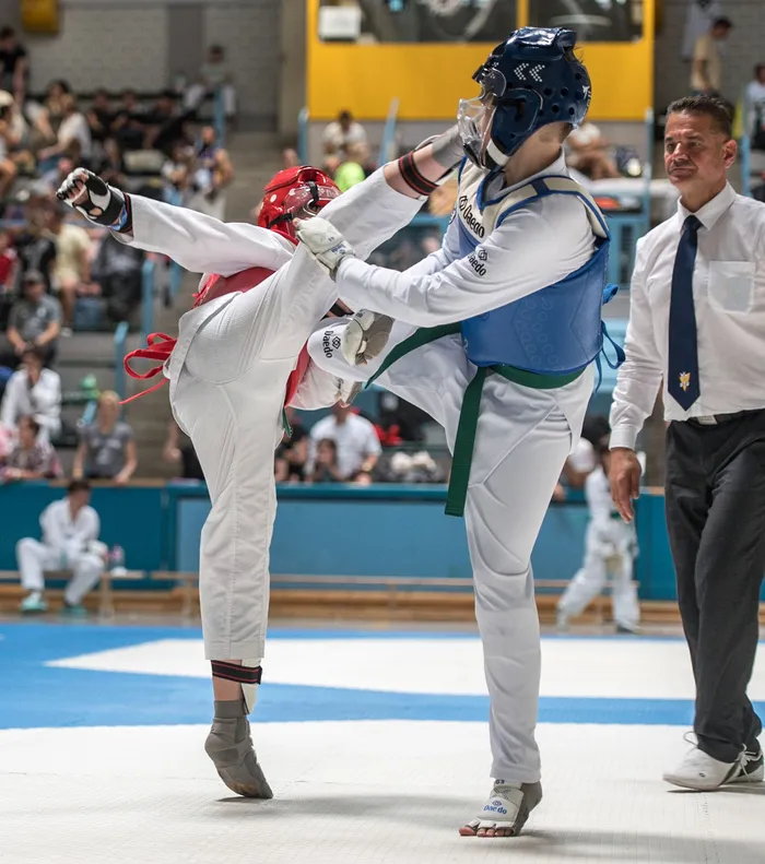 Zwei Taekwondo-Athleten in voller Schutzausrüstung kämpfen in einem Wettkampf. Der Athlet im Vordergrund führt einen Tritt aus, während sein Gegner blockt.