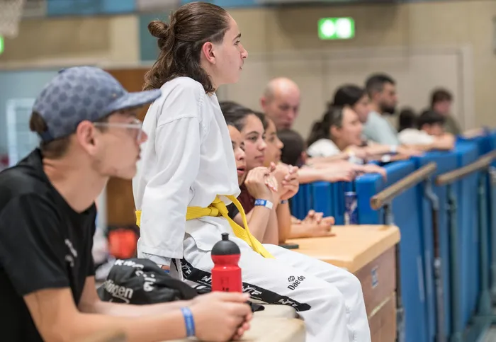 Eine junge Frau mit hochgestecktem Haar und einem gelben Gürtel sitzt in einem weißen Taekwondo-Anzug auf einer blauen Bank und blickt aufmerksam nach vorne. Im Hintergrund sind weitere Personen und eine Sporthalle zu sehen.