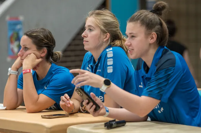 Drei junge Frauen in blauen Trainingsanzügen sitzen an einem Tisch. Eine hält ihr Kinn in der Hand, eine blickt nachdenklich zur Seite, die dritte zeigt auf ihr Smartphone und lächelt.