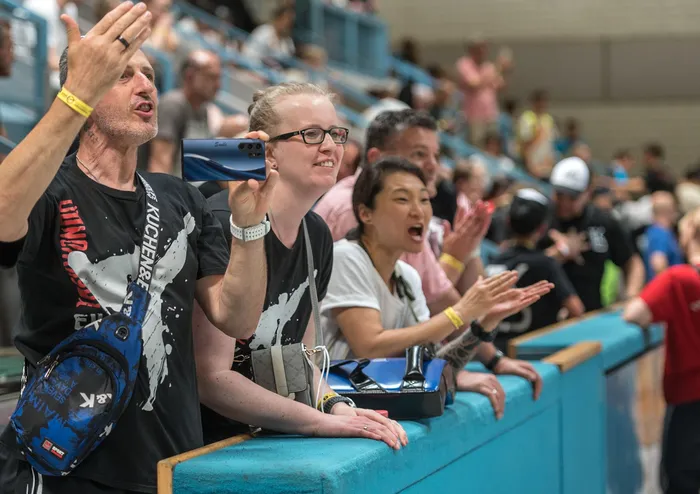Zuschauer in einer Sporthalle applaudieren und filmen mit ihren Smartphones die Kämpfer. Ein Mann im Vordergrund hat die Hände erhoben, eine Frau hält ihr Handy hoch.