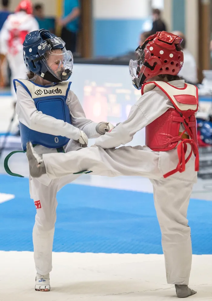 Zwei Taekwondo-Kämpfer in voller Schutzausrüstung treten gegeneinander an. Die Sportlerin in blauem Anzug führt einen Tritt aus. Im Hintergrund sind weitere Sportler und Zuschauer zu sehen.